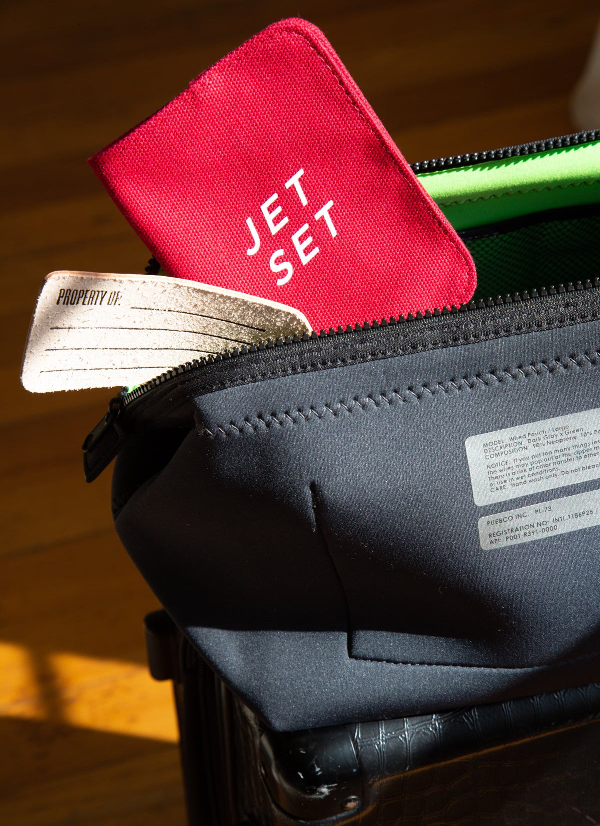 Red 'JET SET' passport holder on top of a black suitcase with a wooden floor background