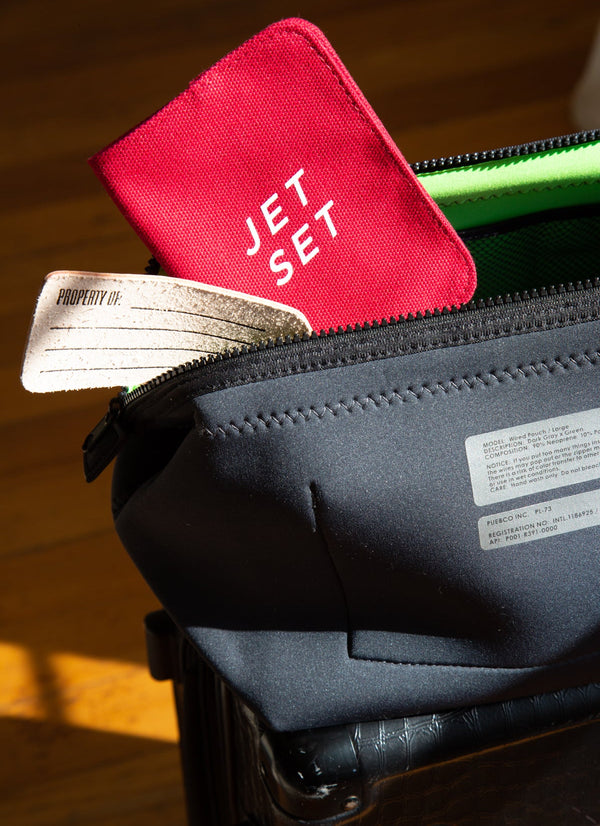 Red 'JET SET' passport holder on top of a black suitcase with a wooden floor background