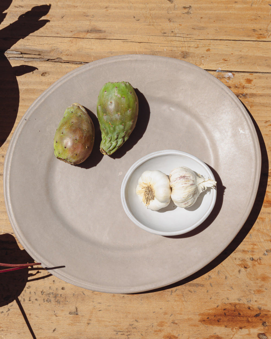 Two garlic bulbs in a small oval bowl on a earth serving platter plate with two green fruits on a wooden surface.