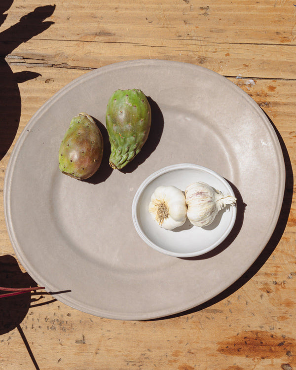 Two garlic bulbs in a small oval bowl on a earth serving platter plate with two green fruits on a wooden surface.