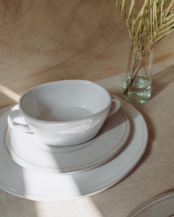 White ceramic bowl and saucer on a wooden surface with a glass of water and plant in the background.